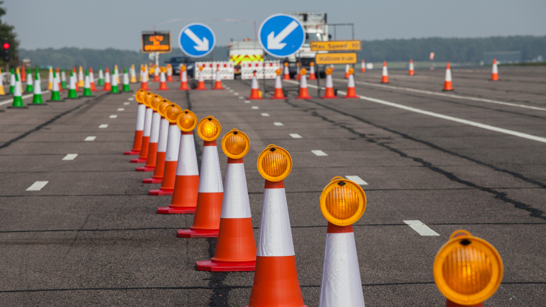 Rectangular image with a daytime shot of 4-lane motorway in the UK closed down to two lanes by a long line of orange and white traffic cones with an orange flashing light on top of each. In the distance, there are two large blue circular signs with white arrows pointing inwards and a blurred announcement sign with orange writing.